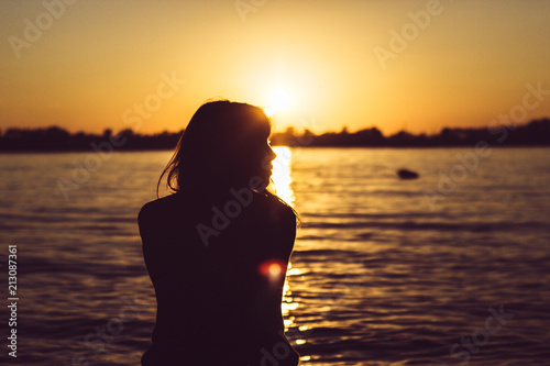 Silhouette of young woman sitting near the water on sand beach with sand in hands © Andrei