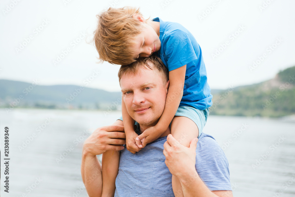 Happy little boy is sitting on his father  On Walk in the summer outdoors. Concept of friendly family..