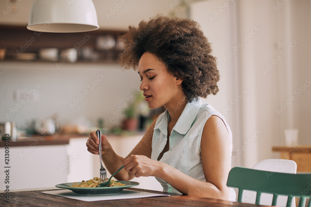Beautiful mixed race woman eating pasta for dinner while sitting at kitchen table. Beautiful mixed race woman eating pasta for dinner while sitting at kitchen table.