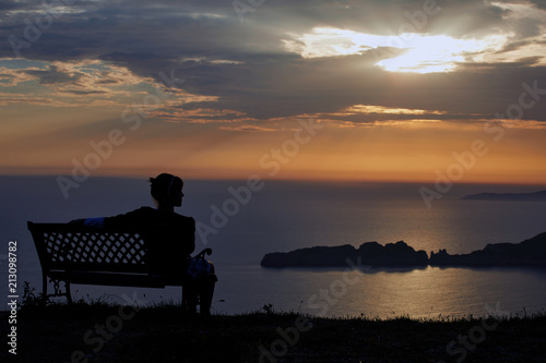 Young woman sits on a bench on a sunset background over the sea and mountains