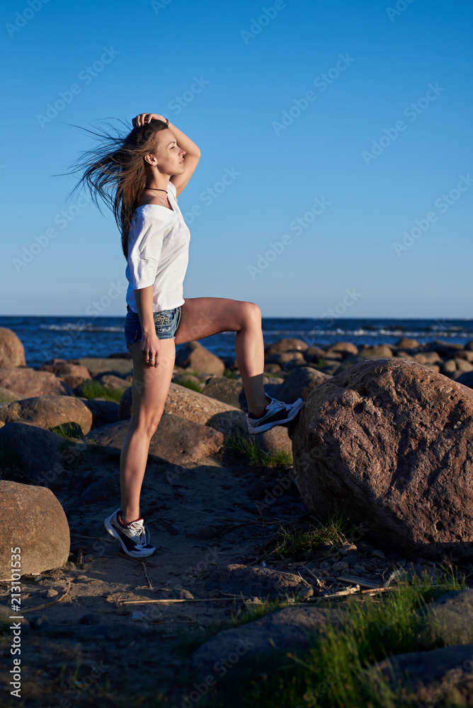 The girl stands on a large stone against the backdrop of rocks and the sea and enjoys the sunset