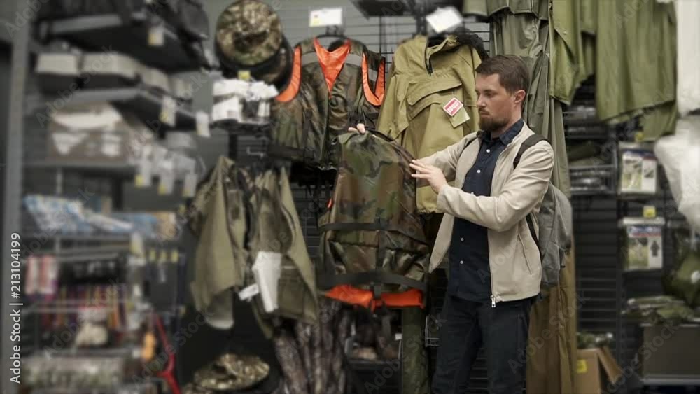 Grown man looking for new lifejacket in sports shop. Male fisher buys new camouflage jacket for fishing. High-quality equipment. Grown man looking for new lifejacket in sports shop. Male fisher buys new camouflage jacket for fishing. High-quality equipment.