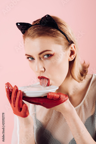 Girl in kitten ears and red gloves with yoghurt on pink background. Dieting and health. Sexy cat woman lick milk from plate. Food and drink. Ecology product and heath. Cat woman eat sour cream.