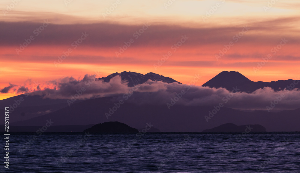 Panoramic photo of mountains behind a lake, hiding in low clouds. Very colorful dramatic sky. Peaceful, relaxing, quiet.