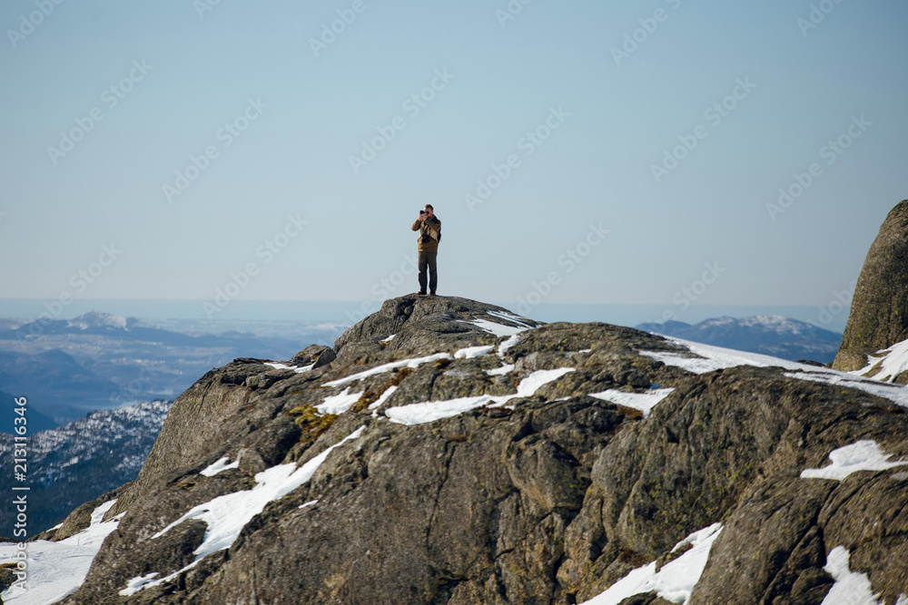 Hiker man makin photo on the camera on the top view of the Pulpit Rock, Preikestolen with beautiful landscape view of sea between rocky shore with snow. Norwegian mountains. Lysefjord, Norway