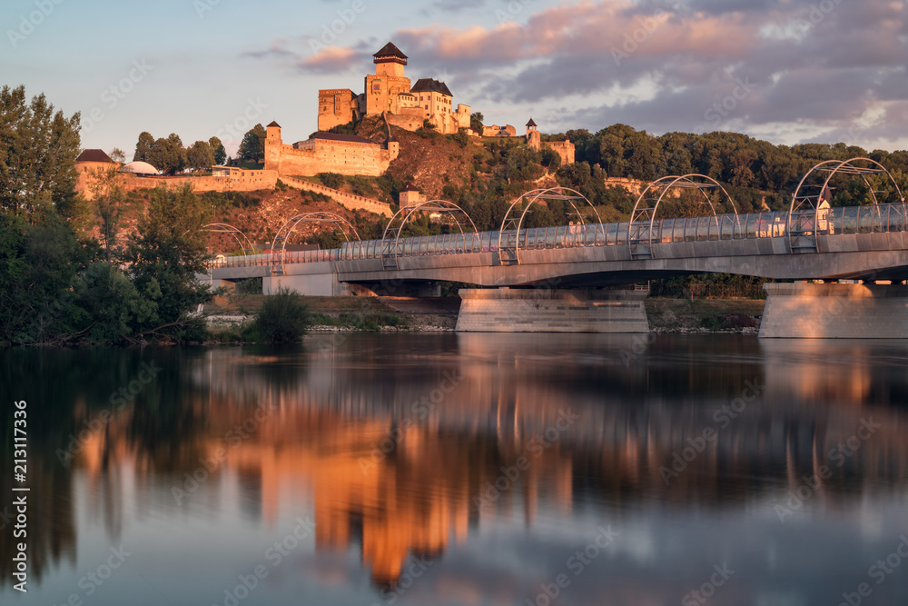 Early meddieval castle of Trencin above a modern railway bridge reflected in a river during a sunset. Trenčianský hrad, Trenčín.