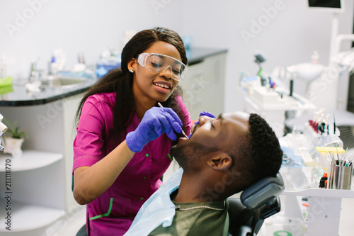 Young african american man lying in chair receiving dental treatment with mouth open, dentist hands wearing gloves holding tools working on patients teeth