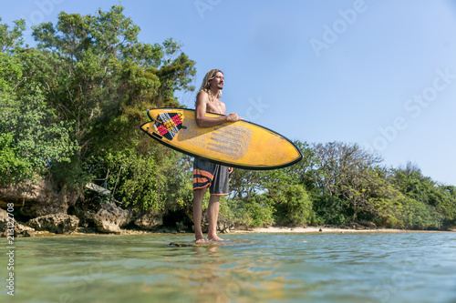 low angle view of shirtless male surfer standing with surfing board in ocean at Nusa Dua Beach, Bali, Indonesia