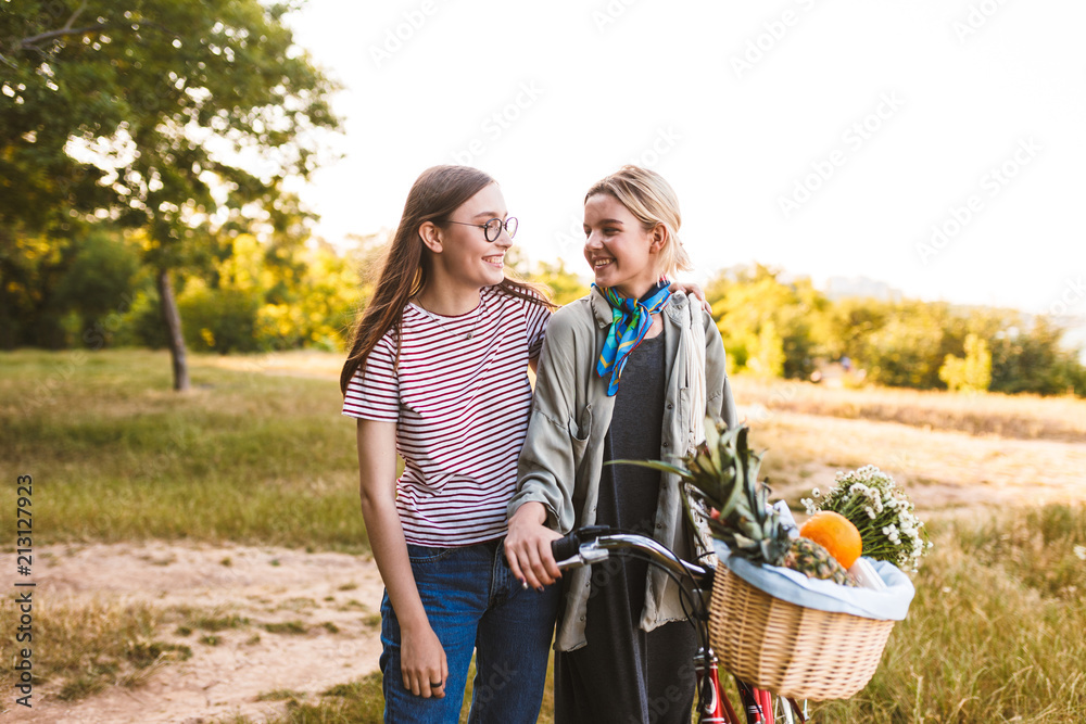 Obraz premium Two beautiful smiling girls with bicycle and basket full of wildflowers and fruits happily looking on each other while spending time in park
