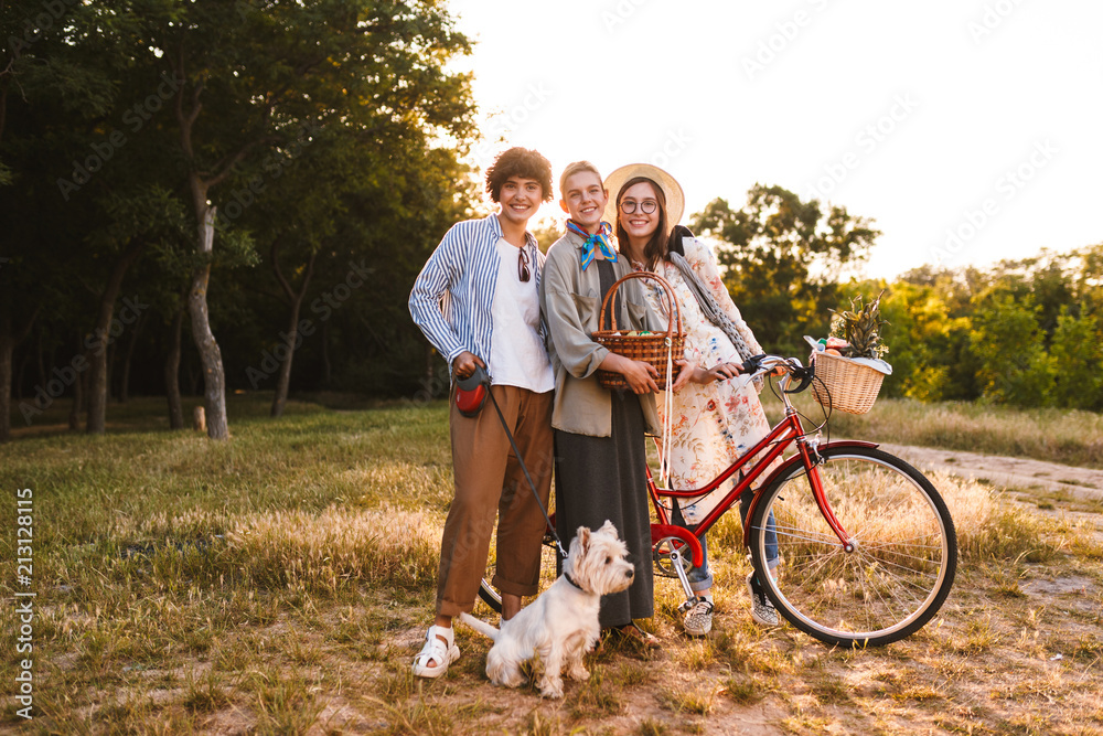 Pretty smiling girls with bicycle and wood basket in hands happily looking in camera while spending time with little dog in park Pretty smiling girls with bicycle and wood basket in hands happily looking in camera while spending time with little dog in park