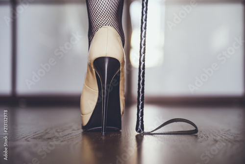 dominant woman in black fishnet stockings and high heels with riding crop, bdsm. Beautiful slim female legs and whip. Strict woman domination