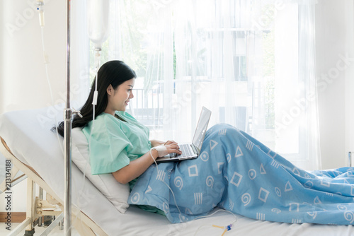 Asian woman sick patient lying in a hospital bed using a laptop for relax when she recovering for ill in hospital..