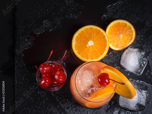 Tequila Sunrise cocktail, orange, ice cubes and maraschino cherries on a wet black slate tray. Top view. photo
