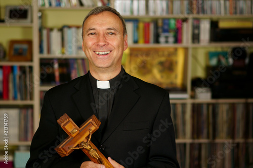 A good looking smiling and laughing catholic priest is showing a crucifix. He looks at us with interest, pensiveness, proselytising, joy and happyness.