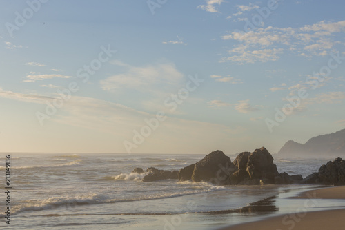 Oregon coast. Rock formation in water with coastal line in background. Hazy air. Low sun.