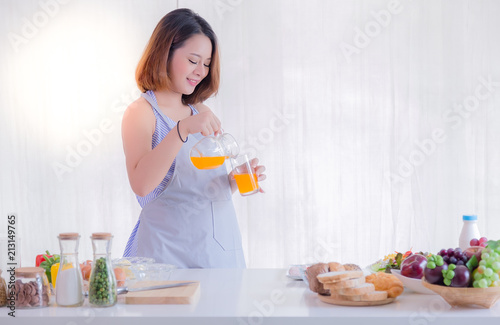 Attractive pregnant woman pours orange juice into the glass. Charming beautiful mother gets thirsty. Gorgeous woman loves drinking juice that makes her smile and happy. She stands in kitchen at home