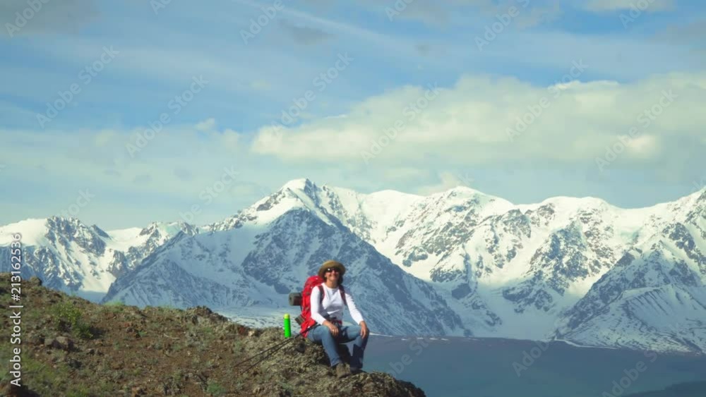 The woman the traveler sits at mountain top, has hot tea from a thermos. Red backpack, orange jacket. Straw hat. Sticks for tracking. The woman the traveler sits at mountain top, has hot tea from a thermos. Red backpack, orange jacket. Straw hat. Sticks for tracking.