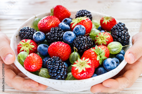 Fresh juicy ripe berries in a white plate in children s hands-strawberries  blueberries  blackberries and gooseberries