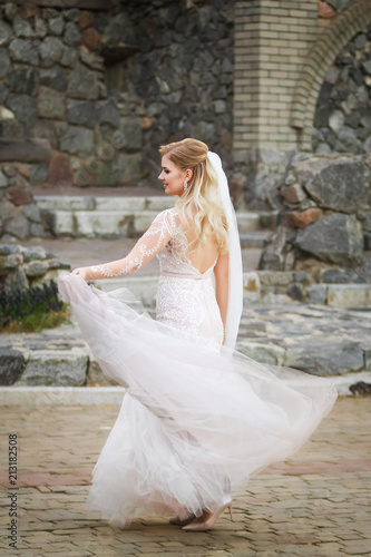 Beautiful sexy smiling bride wearing elegant wedding dress keeping long skirt near the old historical building.fiancee turning backwards and twirling