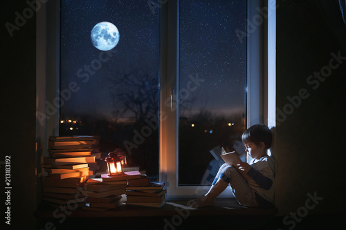 A cute boy sits on the windowsill at night. The child reads books under the moonlight. The window shows the moon and stars.