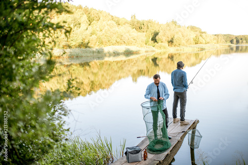 Two male friends preparing for fishing standing with fishing net and rod on the wooden pier during the morning light on the lake