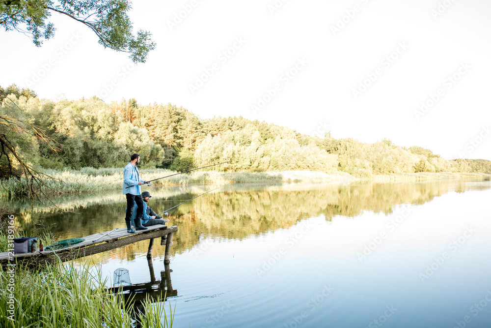 Two male friends fishing together standing on the wooden pier during the morning light on the beautiful lake
