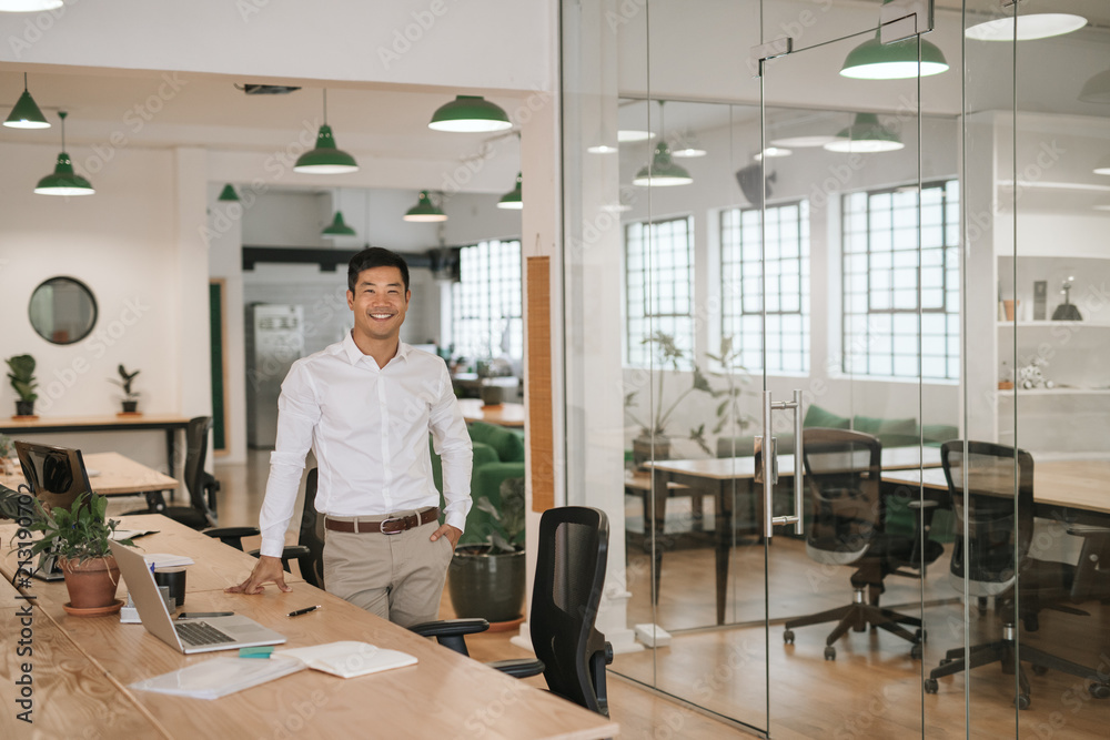 Young Asian businessman smiling while standing by his office desk Young Asian businessman smiling while standing by his office desk