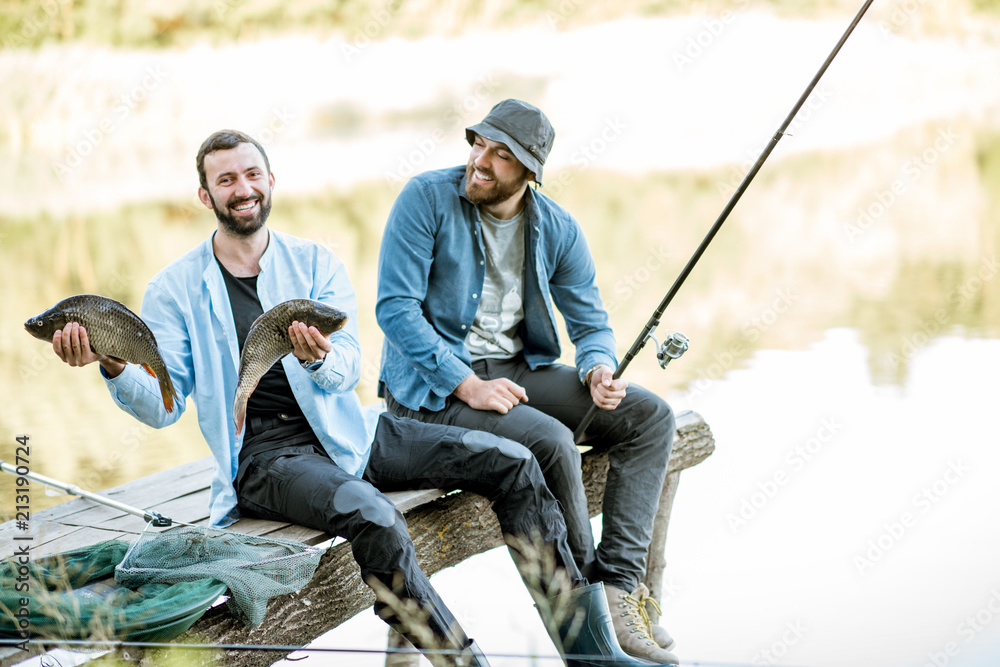Two happy fishermen holding caught fish sitting on the wooden pier during the fishing on the lake at the morning Two happy fishermen holding caught fish sitting on the wooden pier during the fishing on the lake at the morning
