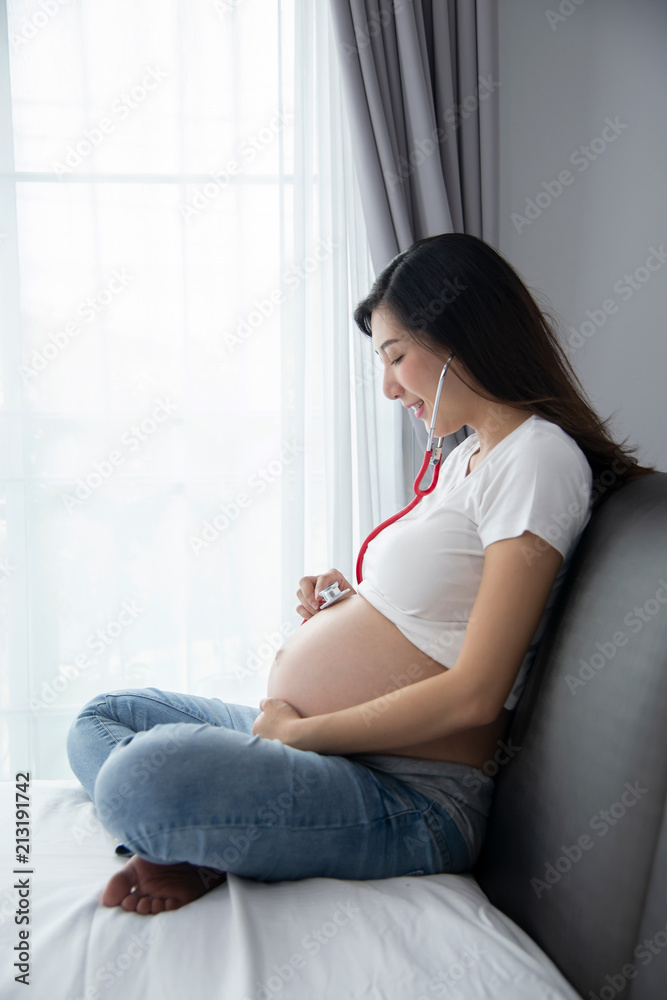Happy Asian pregnant woman using stethoscope in the bedroom.