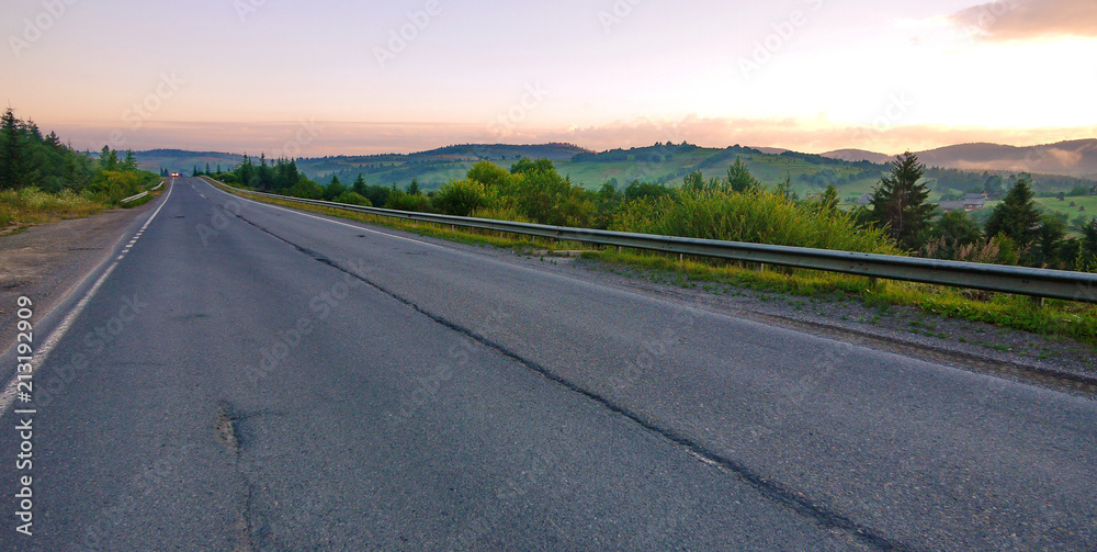 A long straight road running in a mountainous area amidst green plains with a car driving in the distance with lights on. A long straight road running in a mountainous area amidst green plains with a car driving in the distance with lights on.