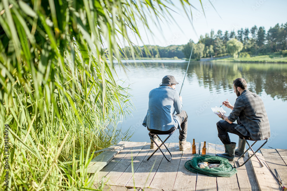Landscape view on the beautiful lake and green reeds with two men fishing on the wooden pier during the morning light Landscape view on the beautiful lake and green reeds with two men fishing on the wooden pier during the morning light