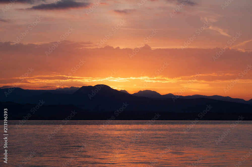 Sunrise Over Lake And Mountains. Early morning landscape. mountain in  silhouettes and the red rays of the rising sun. Sunrise on Lake Garda, Italy