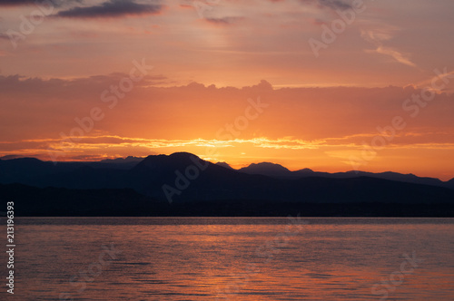 Sunrise Over Lake And Mountains. Early morning landscape. mountain in silhouettes and the red rays of the rising sun. Sunrise on Lake Garda, Italy