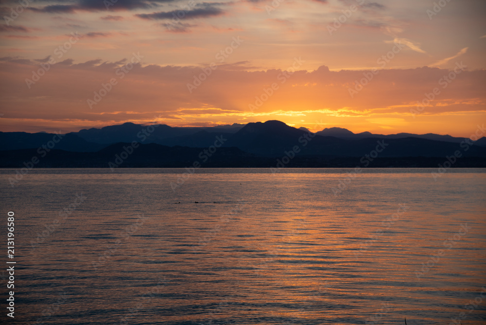 Sunrise Over Lake And Mountains. Early morning landscape. mountain in  silhouettes and the red rays of the rising sun. Sunrise on Lake Garda, Italy