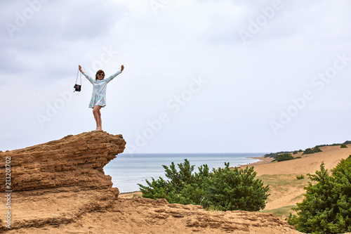 A young woman stands on a rock against a background of blue sea, yellow sand and sky with clouds and admires nature, enjoying solitude