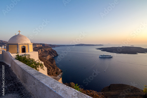 Elevated romantic sunset scene on Santorini. Fira, Greece, from above. Amazing golden hour view from public path walk towards volcano in the caldera. Shortly before the sunset, church in the image
