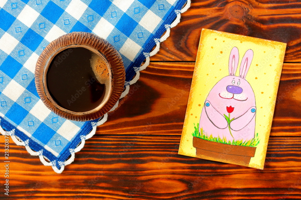 Top view and close up the hot black coffee in a brown ceramic pialat on brown wooden table with blue caged napkin and a postcard with a pink rabbit