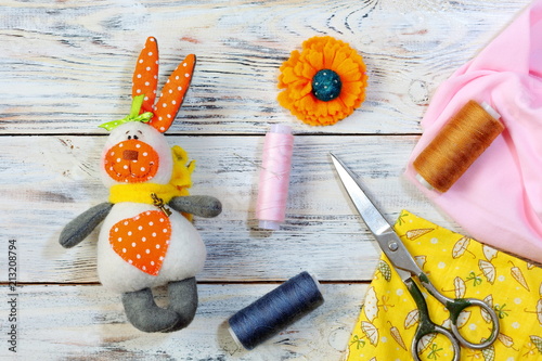 Top view of on a textile bunny toy, cloth scraps, thread reels and scissors. Colorful threads and needles for handicraft lie on white wooden table. The concept of creating textile dolls