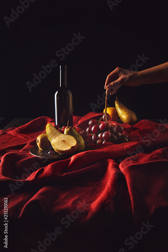 cropped shot of woman taking grapes from table with wine and pears on black photo