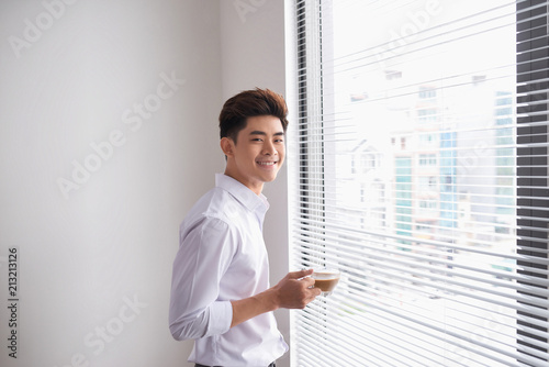 Portrait of a confident young businessman holding a cup of coffee while standing near office window, intelligent men in white shirt while resting after meeting