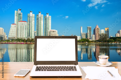 Mockup image of laptop with blank white screen on wooden table view outdoors of office building cityscape background.