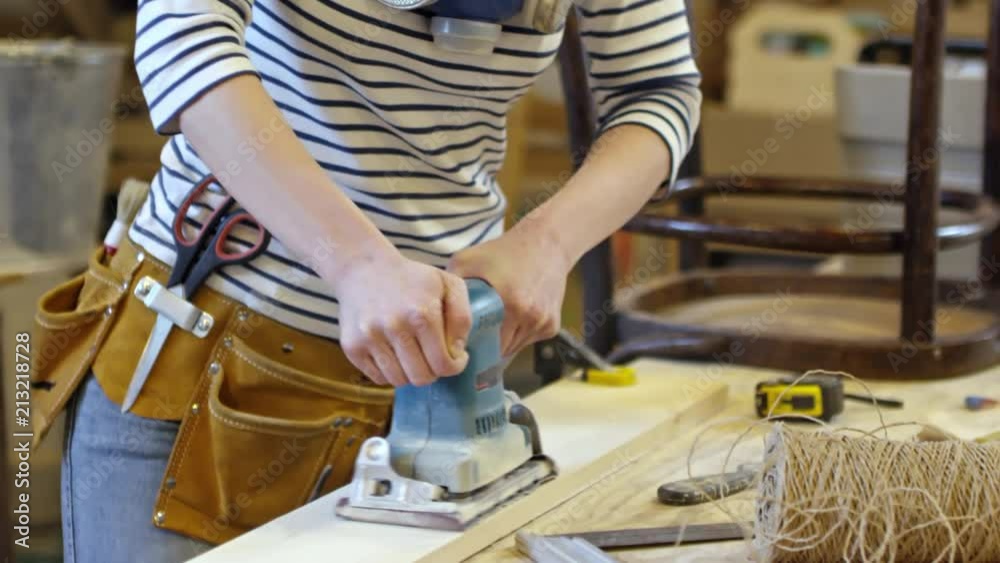 Tilt down shot young woman with respirator polishing wooden plank with sanding machine in carpentry workshop