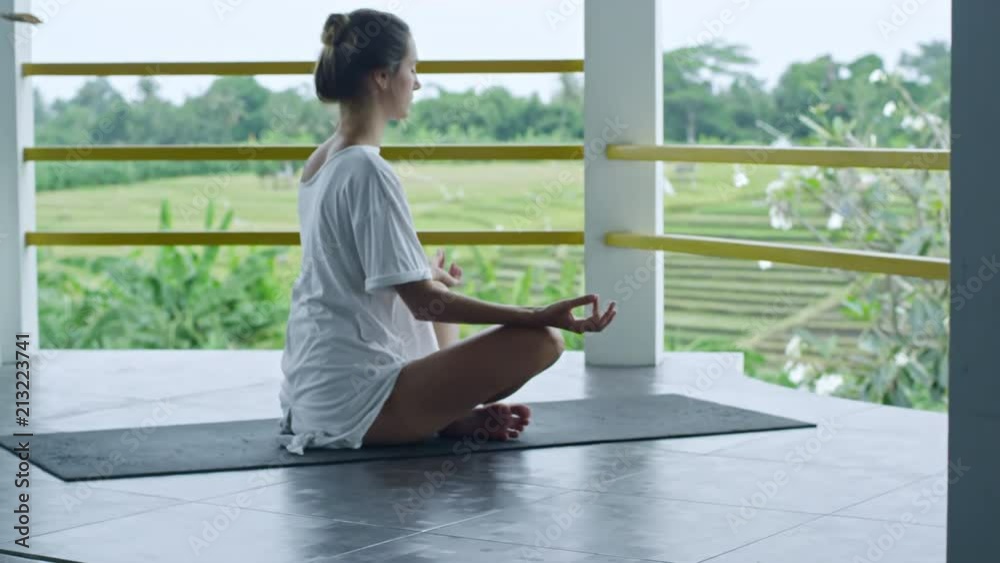 Side view of young woman practicing yoga lotus position when sitting on yoga mat on balcony in the morning Side view of young woman practicing yoga lotus position when sitting on yoga mat on balcony in the morning