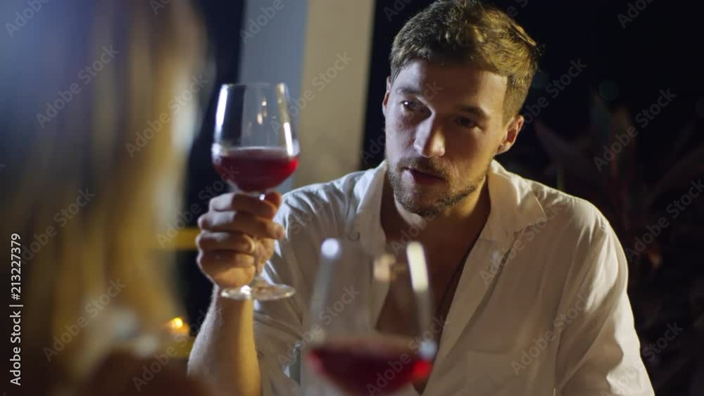Medium shot of handsome man in white shirt drinking wine and smiling when talking to unrecognizable woman seen from her back during date in restaurant Medium shot of handsome man in white shirt drinking wine and smiling when talking to unrecognizable woman seen from her back during date in restaurant