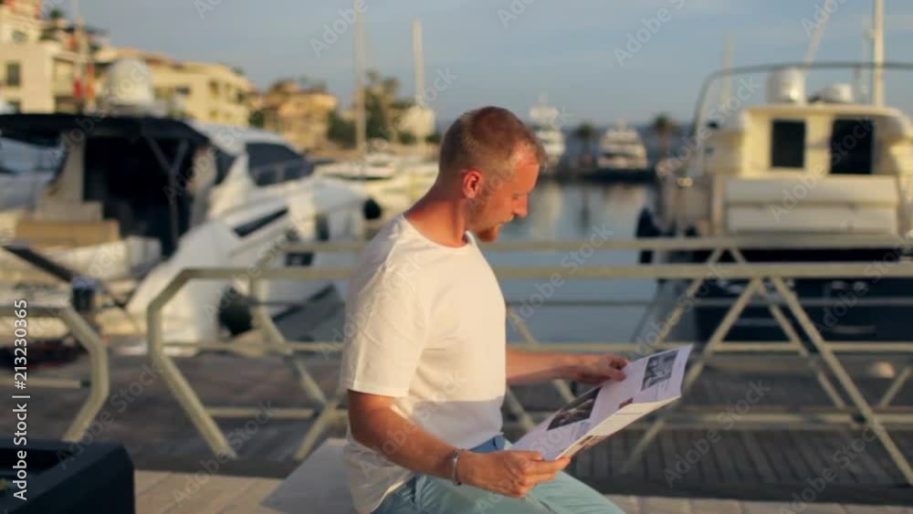 the man is reading a newspaper sitting on the embankment with yachts the man is reading a newspaper sitting on the embankment with yachts