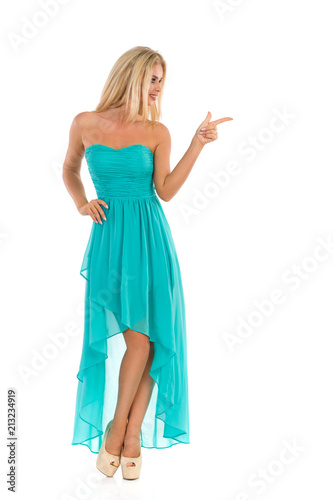 Blond Woman In Turquoise Dress Is Standing, Looking Away And Pointing