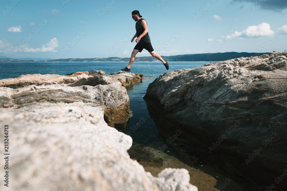 Young tourist man in black casual clothes jumping on the rocks on the stony shore of mediterranean sea in Greece. Sunny summer day