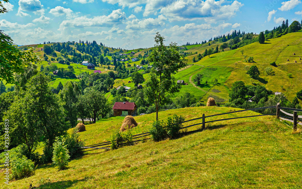 Obraz premium A picturesque plain with huts and haystacks against the background of green mountains and blue sky
