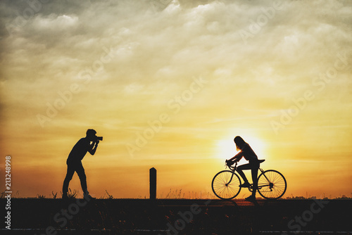 Silhouette of boyfriend taking pictures of girlfriend cyclists. couple are cycling happily in time for sunset.