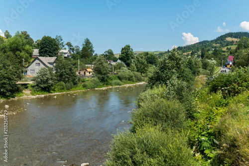 a river flowing along the hidden among the green trees and bushes of private rural houses and forest-covered mountain peaks with a road far away in the distance under a blue cloudy sky. place of rest